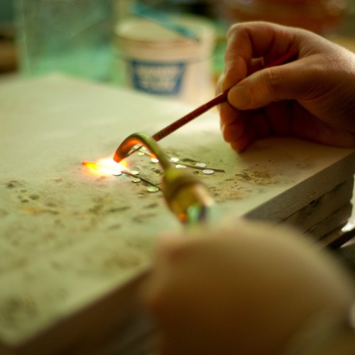A hand holds a tool near glowing metal pieces on a work surface, indicating a metalworking or crafting process.