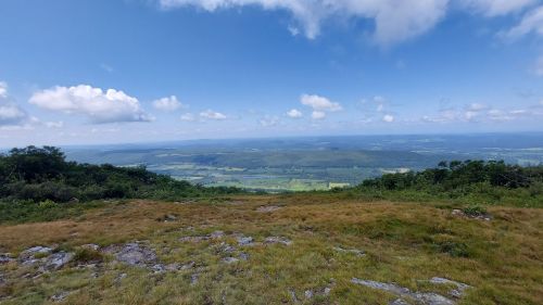 A panoramic view from a mountain summit, showcasing green valleys and a blue sky with scattered clouds.