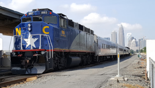 A blue and silver train with "N C" on the front, parked near a city skyline with tall buildings in the background.