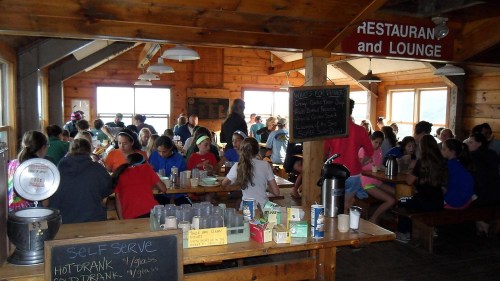 A busy rustic dining hall filled with people enjoying meals at long wooden tables, with a chalkboard menu in view.