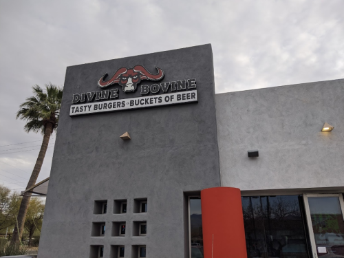 Sign for "Divine Bovine" featuring "Tasty Burgers - Buckets of Beer" on a modern building with palm trees nearby.