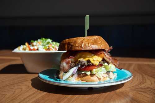 A close-up of a burger with bacon, cheese, lettuce, and tomato on a plate, with a side salad in the background.