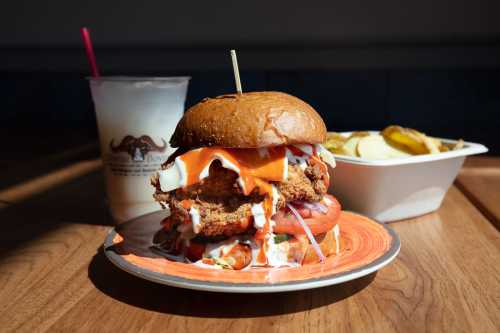 A stacked burger with fried chicken, sauce, and toppings on a plate, alongside a drink and a side of pickles.