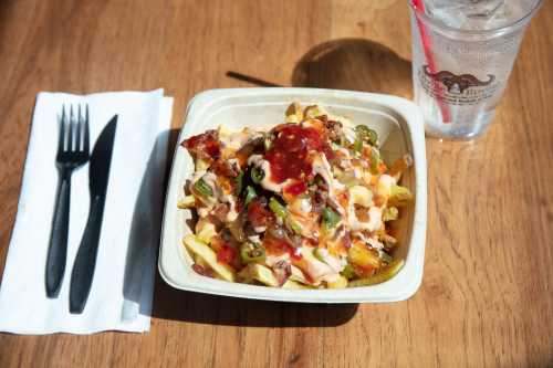 A bowl of loaded fries topped with sauce, jalapeños, and bacon, alongside a fork, knife, and a glass of water.