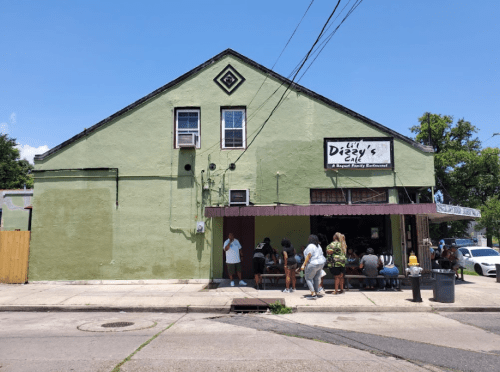 A green building with a sign reading "Dizzy's" and a line of people waiting outside on a sunny day.