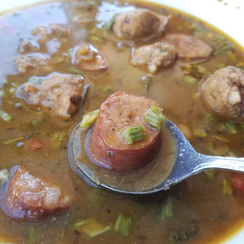 A spoonful of hearty soup with sausage slices and meatballs, garnished with herbs and vegetables.