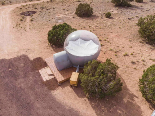 A unique, spherical structure sits on a wooden platform in a desert landscape, surrounded by sparse vegetation.