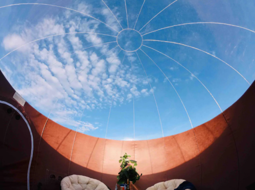 A cozy interior with two chairs and a small plant, featuring a dome-shaped ceiling revealing a blue sky and clouds.