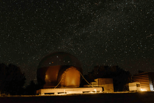 A dome-shaped observatory under a starry night sky, surrounded by trees and faint lights.