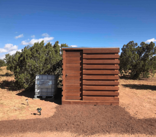 A wooden shed next to a water storage tank, surrounded by sparse vegetation and a clear blue sky.