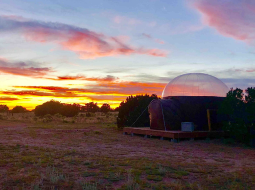 A transparent dome structure sits on a wooden platform against a vibrant sunset sky with scattered clouds.