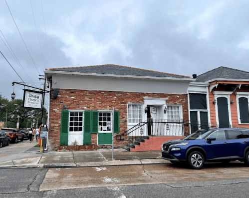 A brick building with green shutters and a sign, located on a street with parked cars and cloudy skies.