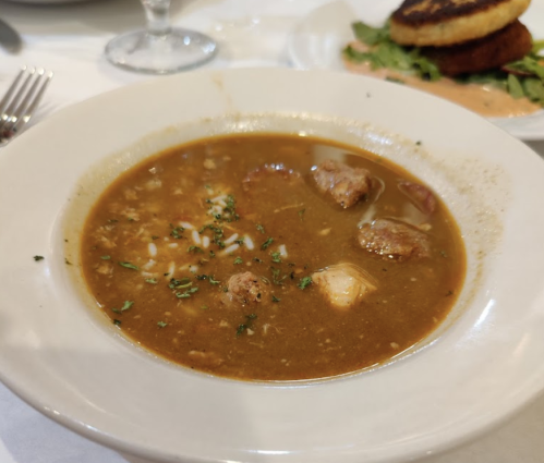 A bowl of hearty soup with meat chunks, herbs, and spices, served on a white plate.