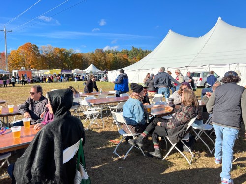 A lively outdoor festival scene with people sitting at tables, enjoying food and drinks, under a clear blue sky.
