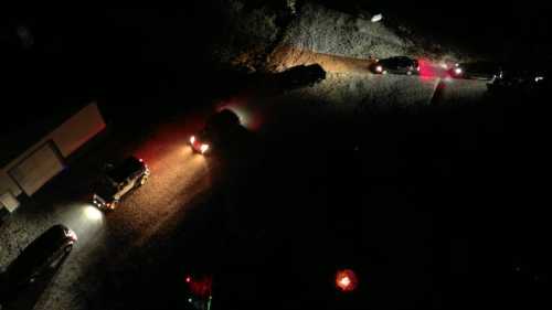 Aerial view of several vehicles with headlights on, parked on a dark, gravel road at night.