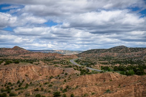 A winding road cuts through a rugged landscape of red rock formations and green vegetation under a cloudy sky.