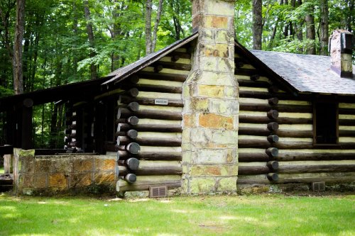 A rustic log cabin surrounded by trees, featuring a stone chimney and a grassy area in front.