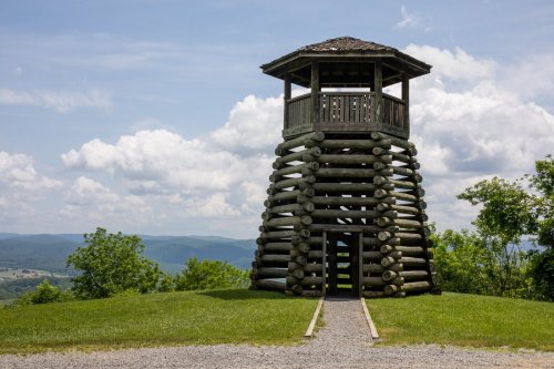 A wooden observation tower with a slatted design, set on a grassy hill with a scenic view of rolling hills and clouds.