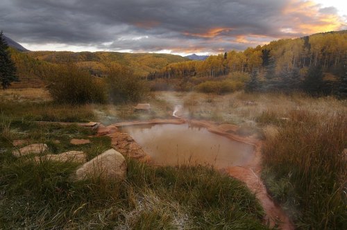 A serene hot spring surrounded by lush grass and trees, with a dramatic sky at sunset in the background.