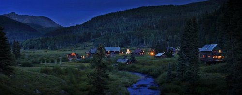 A serene mountain village at dusk, with warm lights glowing from cabins and a stream flowing through lush greenery.