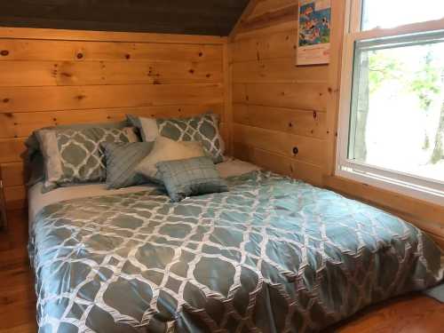 Cozy bedroom with a wooden interior, featuring a bed with teal bedding and decorative pillows, and a window with natural light.