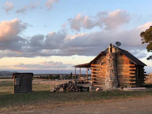 A rustic log cabin with a stone chimney, surrounded by open fields and a cloudy sky at sunset.