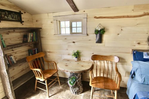 Cozy interior of a small room with wooden walls, two chairs, a round table, and a bookshelf filled with books.
