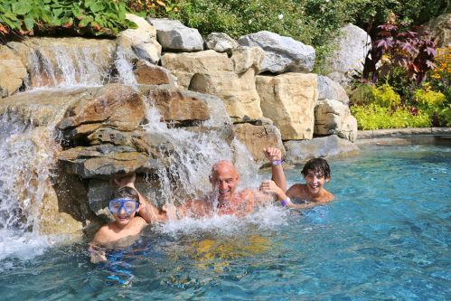 A man and two boys play in a pool with a rock waterfall, enjoying a sunny day.