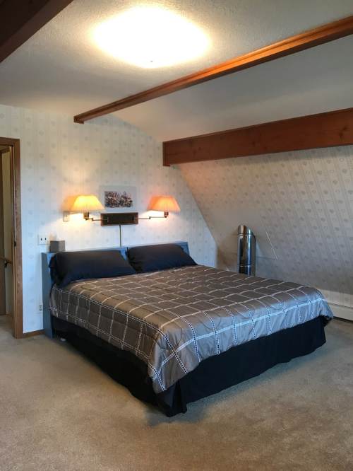 Cozy bedroom with a large bed, two lamps, and a sloped ceiling, featuring neutral decor and carpeted flooring.