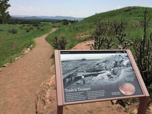 A sign titled "Trash is Treasure" along a scenic trail, with a historical photo and green landscape in the background.