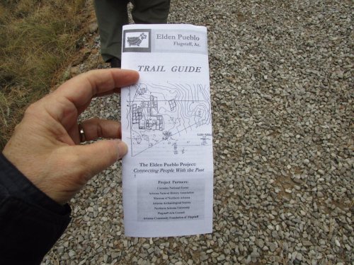 A hand holding a folded trail guide with a map, set against a gravel background.