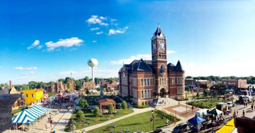 A vibrant town square with a historic clock tower, colorful tents, and people enjoying a sunny day.