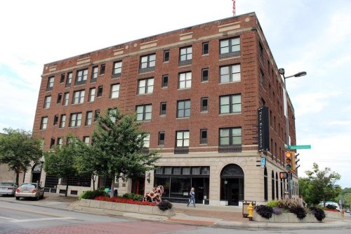 A brick building with multiple windows, surrounded by trees and flowers, on a city street with cars and pedestrians.