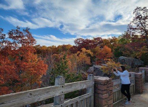 A person takes a photo of vibrant autumn foliage from a wooden overlook against a cloudy sky.