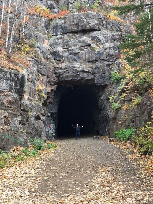 A person stands at the entrance of a dark, rocky tunnel surrounded by autumn foliage and fallen leaves.