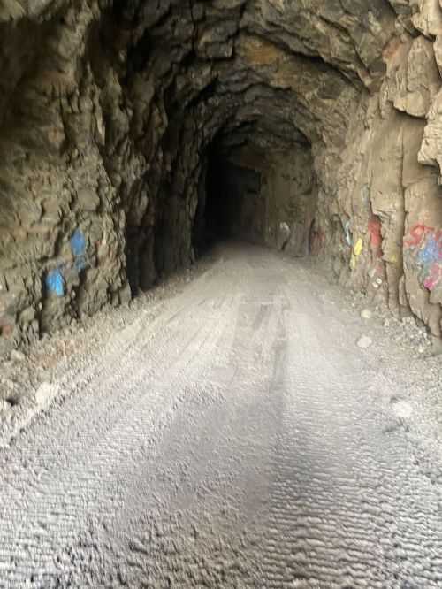 A dimly lit tunnel with rocky walls and a dirt path, featuring colorful graffiti on the sides.