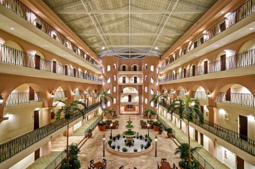 Interior view of a spacious hotel atrium with balconies, palm trees, and a central fountain. Bright and inviting atmosphere.