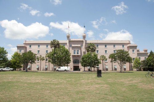 Historic building with a castle-like design, surrounded by trees and a grassy area under a blue sky with clouds.