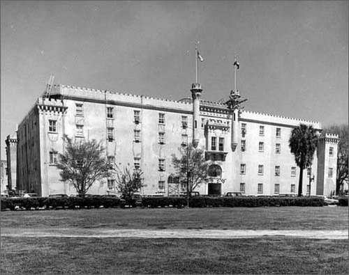 Historic building with a castle-like design, featuring flags and palm trees, set in a grassy area. Black and white photo.