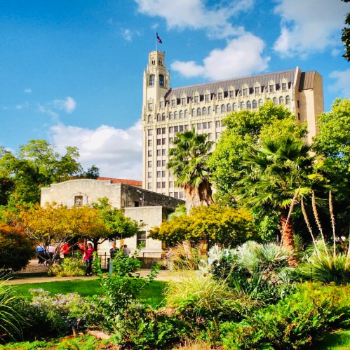 A historic building towers over a lush garden with palm trees and colorful plants under a blue sky with clouds.