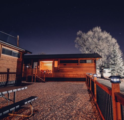 A cozy wooden house at night, illuminated by stars, with a gravel area and trees in the background.