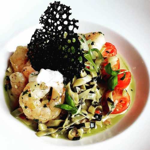 A gourmet dish featuring shrimp, green pasta, cherry tomatoes, and a decorative black crisp on top.