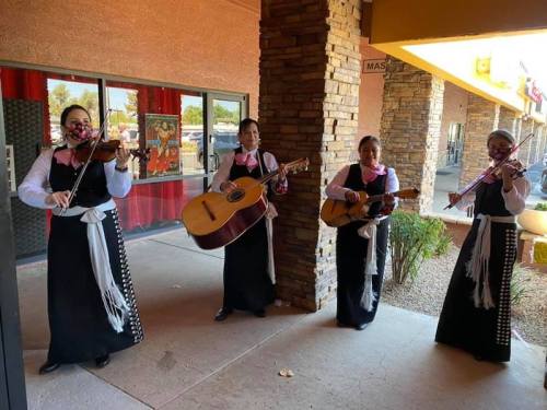 Four women in traditional attire play instruments outside a building, creating a festive atmosphere.