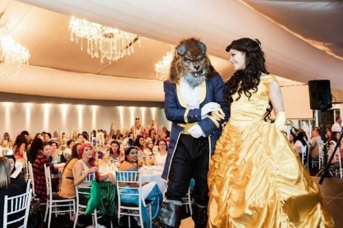 A couple in costume, a lion and a princess, walk through a festive event with guests seated at tables.