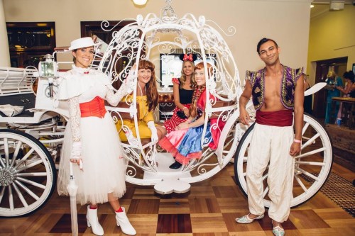 A group of five people in colorful costumes pose together by a decorative white carriage.