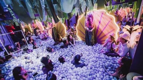 A vibrant party scene with people sitting in a ball pit, surrounded by colorful decorations and dramatic lighting.