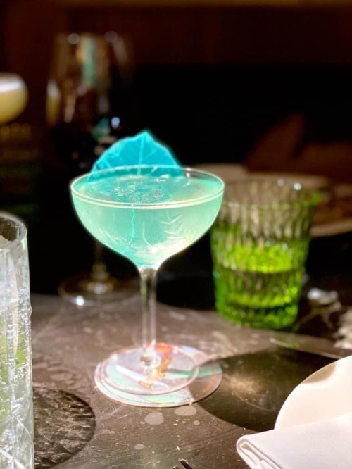 A vibrant blue cocktail in a coupe glass, garnished with a blue leaf, surrounded by green glassware on a table.