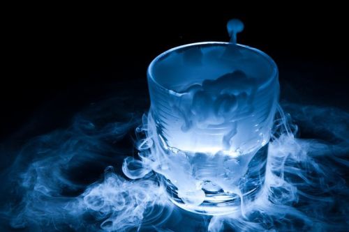 A glass filled with a glowing blue liquid, surrounded by swirling mist or smoke against a dark background.