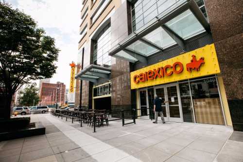 Exterior of Calexico restaurant with a bright yellow sign, outdoor seating, and a modern building backdrop.