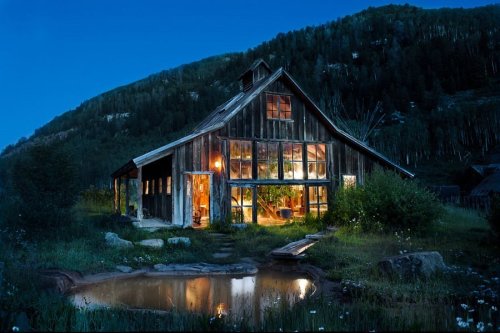 A rustic wooden barn illuminated at night, surrounded by greenery and a small pond, set against a mountainous backdrop.